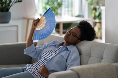 Exhausted African American woman housewife lies on sofa with paper fan after finishing cleaning or other housework. Black girl in casual clothes is stressed because of high temperature in summer clipart