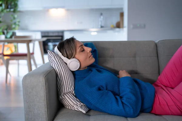 Young woman wearing wireless headphones listens to calm music and closes eyes with satisfaction. Female rests from long hard day lying on sofa in cozy apartment. Girl wearing blue sweater enjoys life