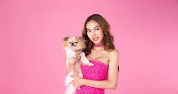 Barbie Girl wear chocky Pink dress hold cute dog and look at camera over pink background. Asian Young woman love puppy and express feeling happy smile, half body portrait, copy space