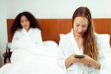 Caucasian and black women love together and lying on bed as diversity person. Young Adult Romantic couple check smart phone behind her back for secret chat text as jealous feeling, copy space