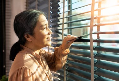 Senior Elderly woman look at window in evening. Grandmother wait patiently for home coming person with sad lonely depress. Asian 70s female waiting with serious worry feeling. copy space