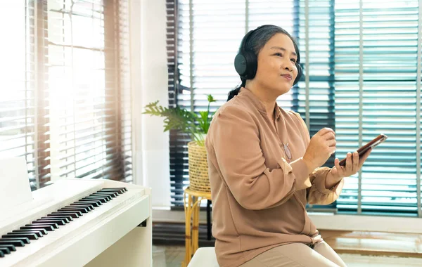 Grandmother listen old music in headphone near piano. Elderly Senior Asian woman enjoy relax from song wireless headset which play oldies songs as think old day and remember cool person, copy space