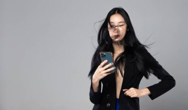 Uzun siyah düz saçlı Asyalı Transseksüel Kadın. Rüzgâr gibi esiyor. Kadın telefonu rüzgar fırtınasına karşı tutuyor, moda tutkusu seksi, gri arka plan izole edilmiş fotokopi alanı