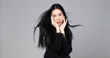 Uzun siyah düz saçlı Asyalı Transseksüel Kadın. Rüzgâr gibi esiyor. Kadın telefonu rüzgar fırtınasına karşı tutuyor, moda tutkusu seksi, gri arka plan izole edilmiş fotokopi alanı