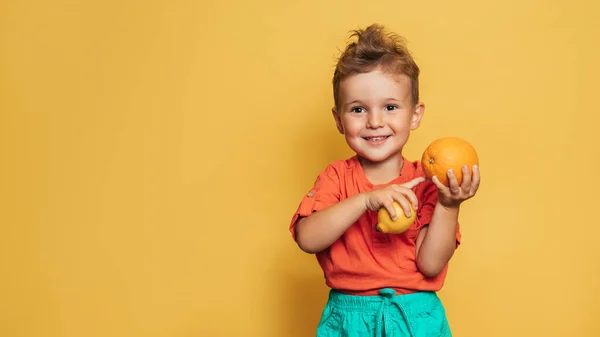 Sarı arka planda taze limon ve portakal tutan gülümseyen bir çocuğun stüdyo fotoğrafı. Sağlıklı bebek maması konsepti, C vitamini metnin için bir yer..