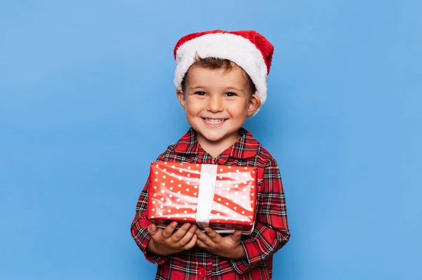 Een gelukkig jongetje in een kerstpyjama en een hoed met een geschenkdoos. De studio opname is verguld op een blauwe achtergrond. Het begrip vakantie. — Stockfoto