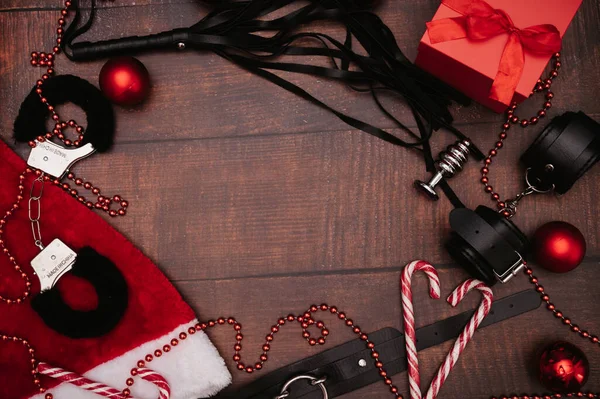 Noel dekorlu bir çift yetişkin BDSM oyuncağı. Metin için alanı olan bir çerçeve. Düz çizgi. Bir armağan örneği. Kelepçe, kırbaç, anal tıkaç, Noel topları..
