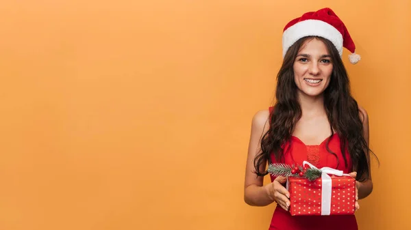 Uma mulher branca sorridente em um chapéu vermelho de Papai Noel com uma caixa de presente em suas mãos em um fundo amarelo isolado. O conceito de Natal e Ano Novo. Um lugar para o seu texto. — Fotografia de Stock