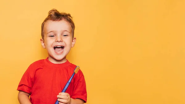 O garoto feliz está segurando uma escova de dentes azul em um fundo amarelo e sorrindo mostrando seus dentes. Saúde, higiene oral. Um lugar para o seu texto. — Fotografia de Stock