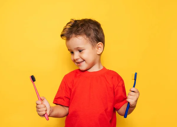 O garoto feliz segura 2 escovas de dentes em um fundo amarelo em ambas as mãos e sorri mostrando seus dentes. Saúde, higiene oral — Fotografia de Stock