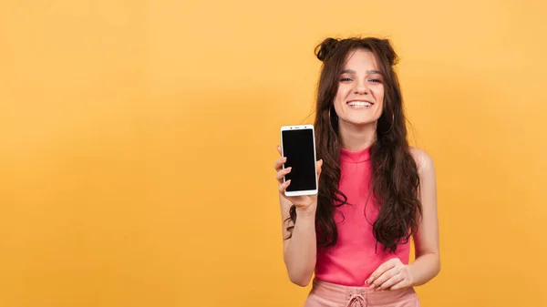 Uma mulher sorridente segura um smartphone com uma tela clara, em pé sobre um fundo amarelo. — Fotografia de Stock