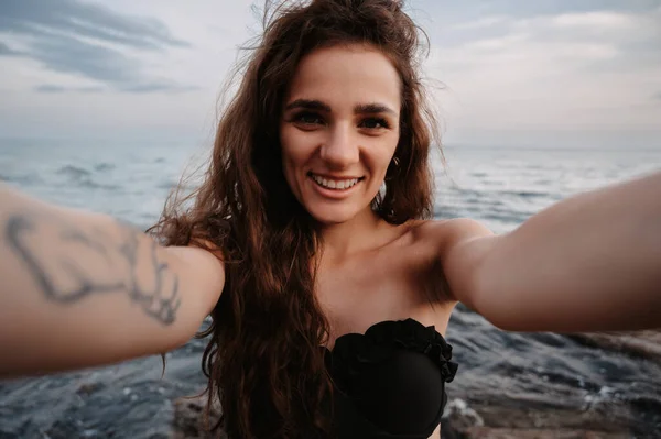 Selfie-portrait of an attractive girl with long hair smiling into the camera close-up, standing against the background of the sea. The concept of tourism, resort, vacation. — Foto Stock