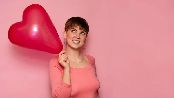 Portrait of a bright young woman with a red heart-shaped balloon on a pink background. A place for your text. — Stok fotoğraf