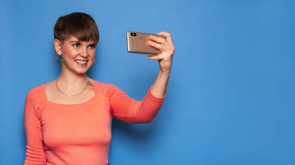 Retrato de uma jovem mulher tirando uma selfie em um smartphone em um fundo azul. Um lugar para a tua mensagem. O conceito de comunicação por vídeo à distância — Fotografia de Stock