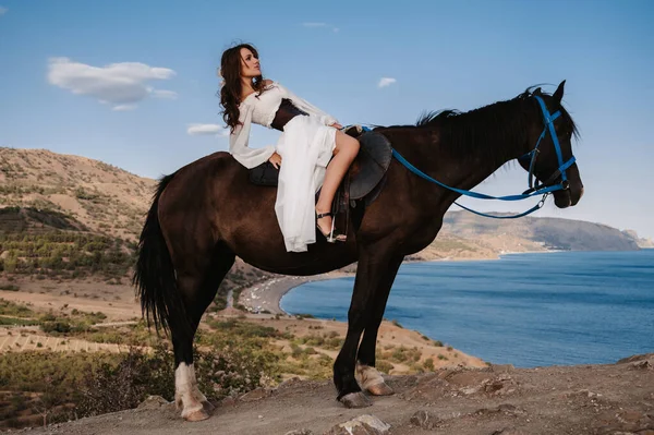 Une jeune cavalière en robe blanche pose en selle sur un cheval appuyé contre un beau paysage de montagne. Le concept de romance, de contes de fées. Exemple de couverture de livre fini — Photo