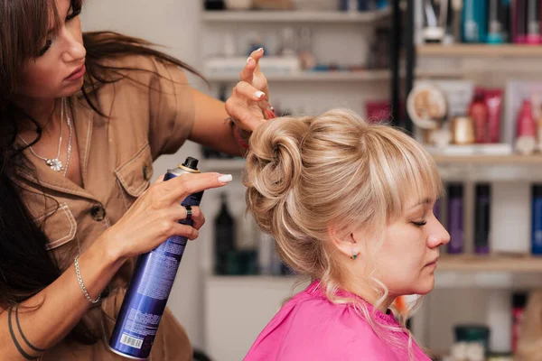 A menina-cabeleireiro faz o cliente um penteado de uma celebração. O cabeleireiro corrige tudo com spray de cabelo — Fotografia de Stock