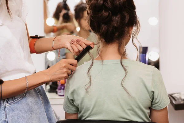 A filmar num salão de beleza. O mestre do cabelo corrige o penteado de uma jovem garota de cabelos escuros com a ajuda de uma escova de cabelo. — Fotografia de Stock