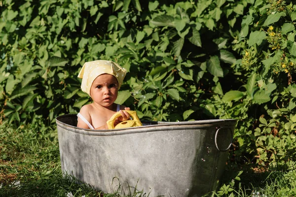 Una niña pequeña en un pañuelo para la cabeza y con una regadera se está bañando en el baño de los niños en el jardín, en un día soleado caliente del verano —  Fotos de Stock