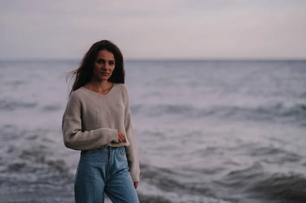 A young Caucasian girl stands on a rocky beach by the sea, with a thoughtful look. Gray, cool, inclement weather. — Stock Photo, Image