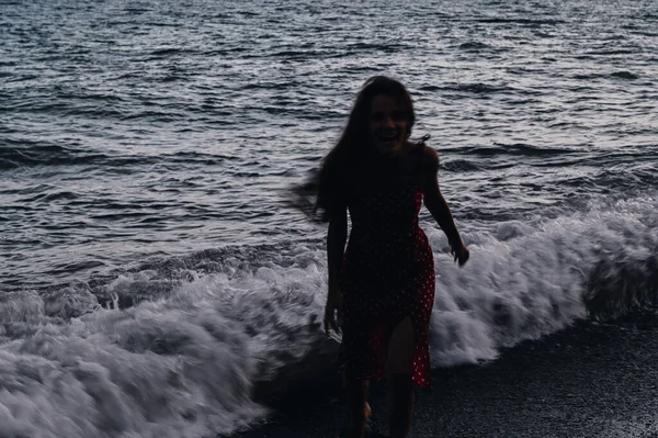 夏の夜、波の上を海岸線に沿ってドレス姿の少女が走っている。自由の概念. — ストック写真
