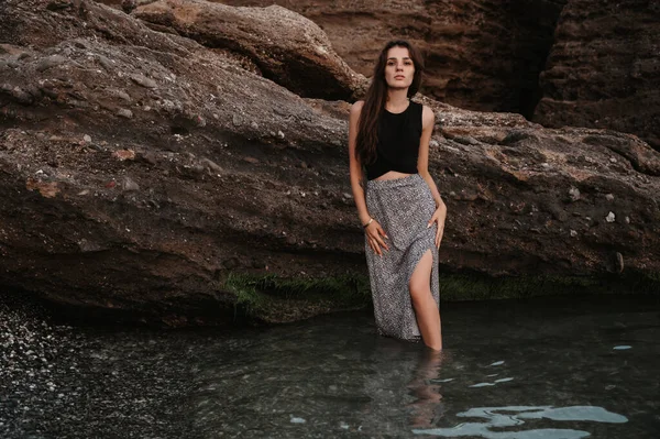 Une photo d'une belle jeune femme sensuelle aux longs cheveux foncés dans une robe posant sur la plage par de grands rochers. — Photo