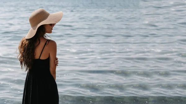 ドレスとわらの帽子で長い暗い髪を持つ美しい若い女性の写真は、ビーチにポーズをとって、彼女の背中と立って無限の海を見ています。テキストの場所. — ストック写真