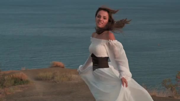 En ung vacker flicka med mörkt hår, klädd i en vit klänning, snurrar på toppen av berget, inför vinden blåser hennes hår och klänning, och ler - harmoni, äventyr, frihet, 4k video — Stockvideo