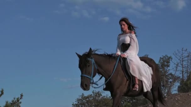 Eine junge Reiterin im weißen Kleid steigt hoch zu Ross vor der Kulisse einer wunderschönen Berglandschaft vom Hang ab. Abenteuer, Freiheit, 4k video, Zeitlupe — Stockvideo