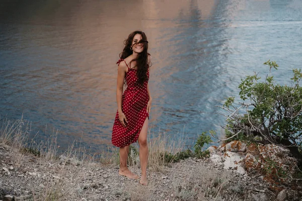 Une photo d'une jeune femme caucasienne en robe rouge au bord d'une falaise près de la plage et de la mer avec un sourire sincère sur son visage et des cheveux emmêlés par le vent. Le concept de liberté et de voyage — Photo