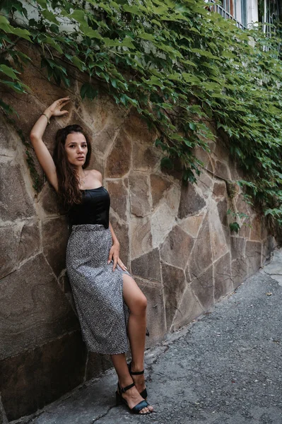 Una giovane bruna posa contro un muro di pietra con edera verde. Una figura bella e snella. Una ragazza con un vestito. — Foto Stock