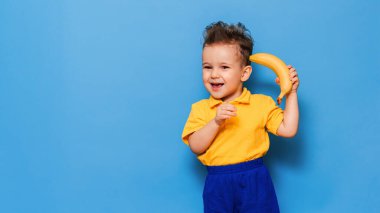 Muzlu komik surat. Sarı tişörtlü bir çocuk elinde gülümsemeli bir muz tutuyor. Komik ifadeler. Beslenme kavramı. Taze muz. Sarı stil. Mavi arkaplan