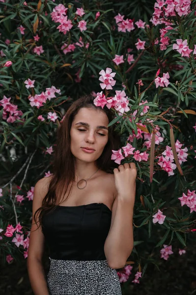 Retrato de una joven soñadora de una hermosa morena en un jardín floreciente de adelfa rosa. Concepto de estilo de vida en viajes de verano —  Fotos de Stock