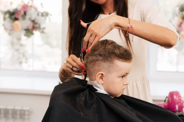 A filmar num salão de beleza. Um barbeiro corta um cabelo de garotinho com tesoura — Fotografia de Stock