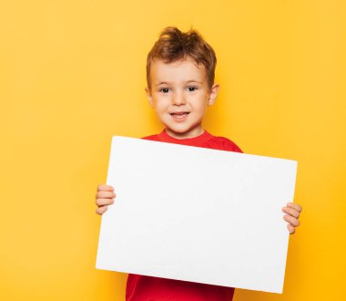 Studio portrait of a happy boy with a clean white poster in his hands on a bright yellow background, with a place for your text or advertising