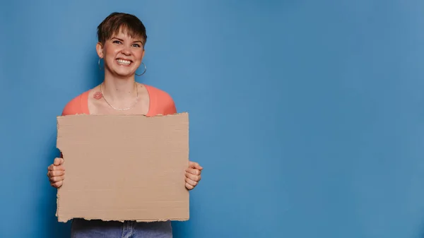 Mavi arka planda boş bir karton çarşaf tutan gülümseyen genç bir kadının stüdyo fotoğrafı. İlanınız veya metniniz için boşluk kopyala. — Stok fotoğraf