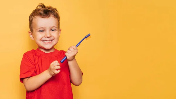 O garoto feliz está segurando uma escova de dentes azul em um fundo amarelo e sorrindo mostrando seus dentes. Saúde, higiene oral. Um lugar para o seu texto. — Fotografia de Stock