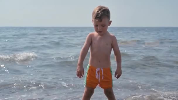 A cute baby boy is standing in the sea on the coast. Summer holidays and sea holidays. Authentic, slow-motion video — Vídeo de Stock