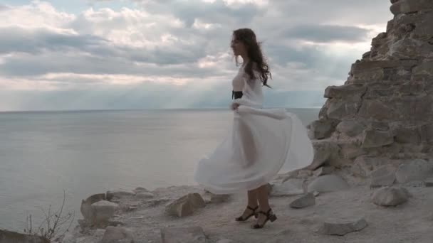 A young girl with dark hair, dressed in a white dress, is spinning on the top of the mountain, the wind blowing her hair and dress, and smiling - harmony, adventure, freedom, 4k video. slow motion — Stockvideo