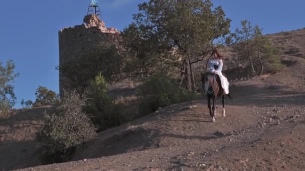 Eine junge Reiterin im weißen Kleid steigt hoch zu Ross vor der Kulisse einer wunderschönen Berglandschaft vom Hang ab. Abenteuer, Freiheit, 4k video, Zeitlupe — Stockvideo