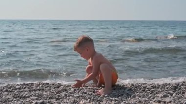 Cute baby boy playing with stones on the beach, on the seashore. Summer holidays and sea holidays. Authentic, slow-motion video
