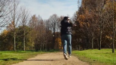 Bir kız parkın sonbahar sokağında koşuyor. Kot pantolon ve ceket giymiş genç bir kadın. Altın yapraklar yerde yatıyor.
