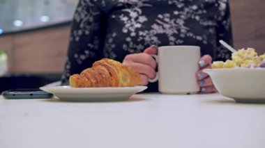 Masadaki bir kadın kahve içiyor. Bir kruvasanın yakın plan çekiminde, bir tabakta. Kızın elleri bir kupa tutuyor.