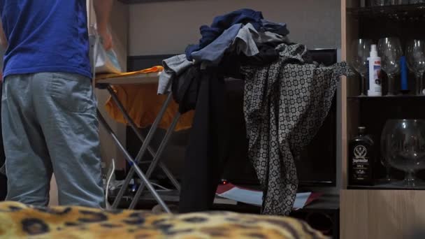 Un chico planchando una camiseta con una plancha. tareas domésticas. Hay una pila de ropa arrugada en la tabla de planchar. vista desde atrás — Vídeos de Stock