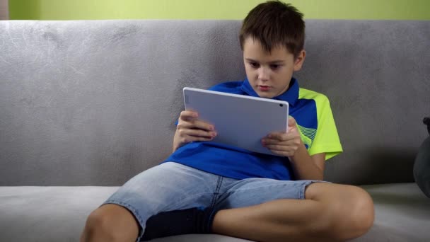 Подросток сидит за планшетом. с энтузиазмом играть. мальчик в футболке и шортах сидит на диване в комнате — стоковое видео