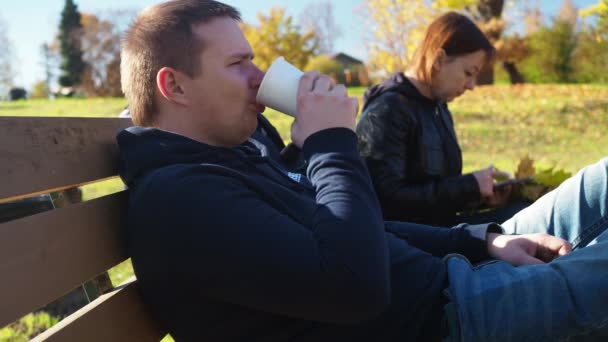 Egy férfi és egy nő ül egy padon a parkban. Egy nő épp egy kazettát lapoz át a telefonján, egy férfi lassan teázik egy bögréből. családi nyaralás a természetben — Stock videók