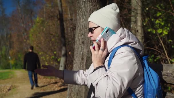 Uma mulher idosa está sentada em um banco de estacionamento e falando ao telefone em um caso. Outono no parque. ela está usando um casaco, um chapéu e óculos de sol. close-up. as pessoas passam — Vídeo de Stock
