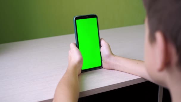 Looks at the touch screen. a teenager is sitting with a green-screen phone at a table in the room. the face is not visible — Stock Video