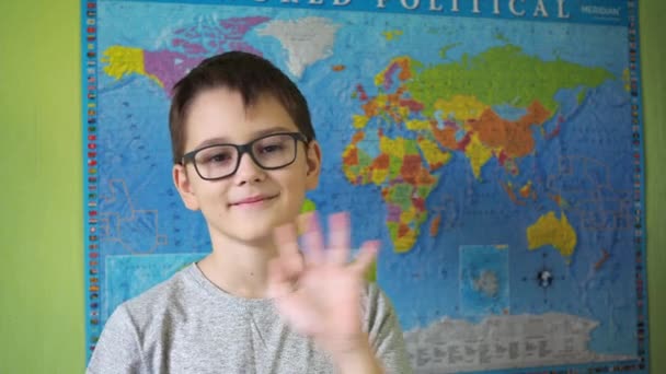 Un niño con gafas agita su mano de pie sobre el fondo de un mapa político del mundo. una sonrisa en la cara de un adolescente — Vídeo de stock