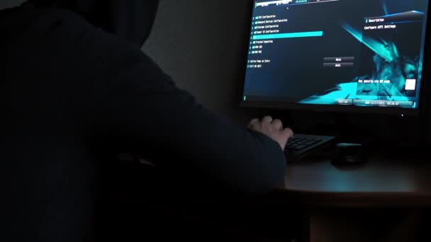 Een man in een sweatshirt met capuchon zit achter een computer in het donker. licht van de computer. het gezicht is niet zichtbaar. programmering — Stockvideo