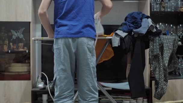 Un chico planchando una camiseta con una plancha. tareas domésticas. Hay una pila de ropa arrugada en la tabla de planchar. vista desde atrás. sin cabeza — Vídeos de Stock